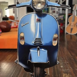 May include: A light blue Vespa scooter with a glossy finish. The scooter has a round headlight, side mirrors, and the Vespa logo. The scooter is displayed on a wooden floor.