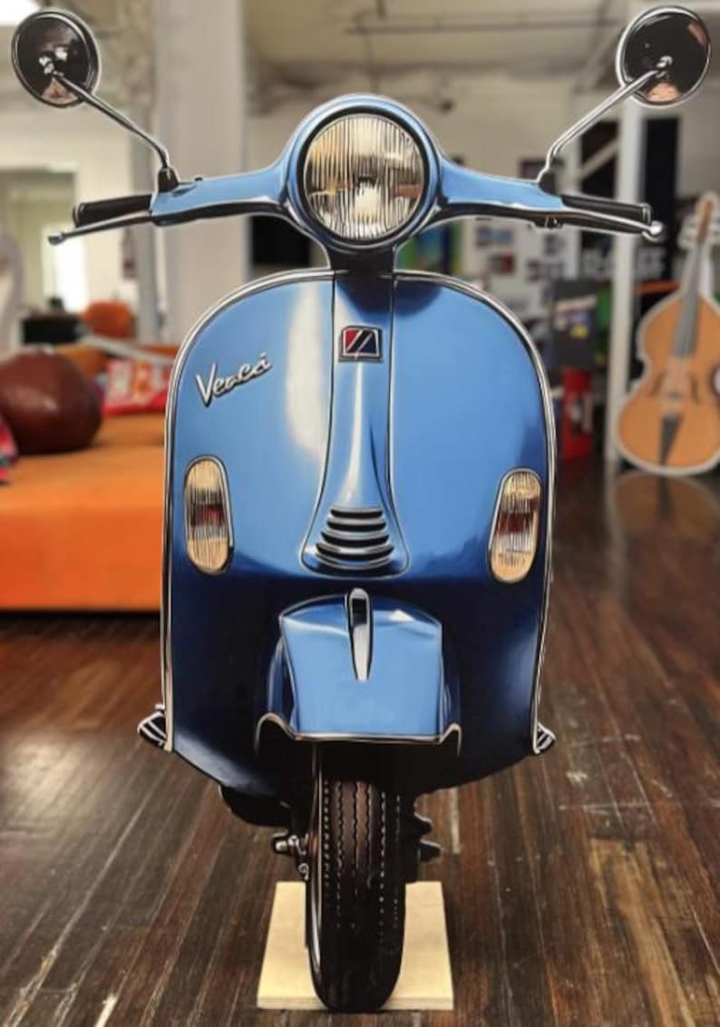 May include: A light blue Vespa scooter with a glossy finish. The scooter has a round headlight, side mirrors, and the Vespa logo. The scooter is displayed on a wooden floor.