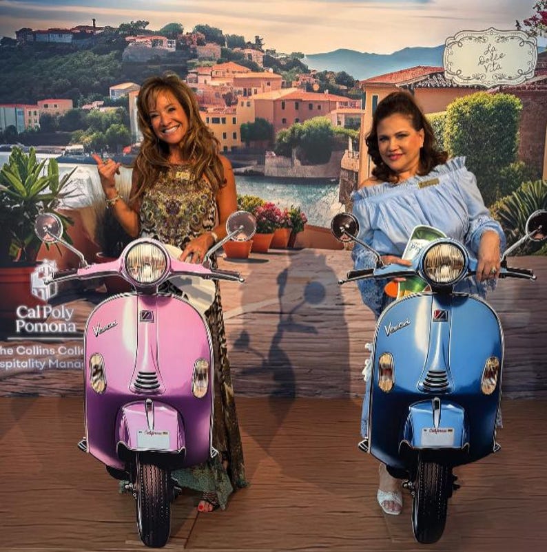 May include: Two women stand with classic scooters. One is pink and purple, the other blue. The scooters have chrome details and headlights. The backdrop is a mural of a coastal town, evoking a sense of travel.