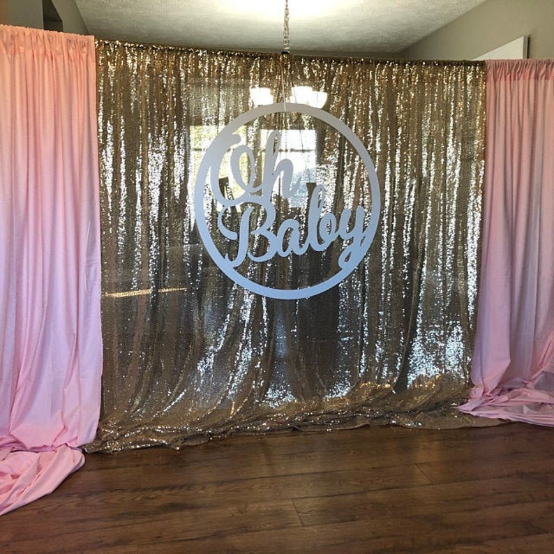 May include: A gold sequin backdrop with pink curtains on either side. A white circle with the text "Oh Baby" hangs in the center of the backdrop.