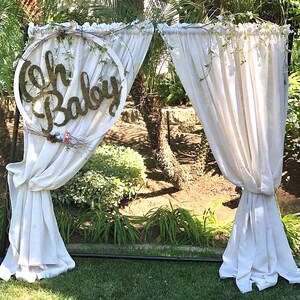 May include: A white fabric backdrop with a wooden sign that says "Oh Baby" in a circle. The backdrop is set up outdoors with greenery in the background.