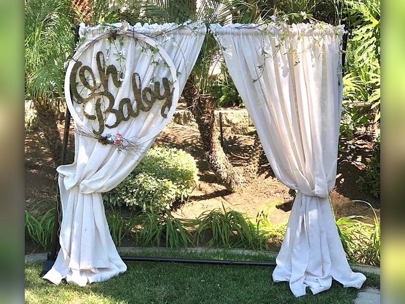 May include: A white fabric backdrop with a wooden sign that says "Oh Baby" in a circle. The backdrop is set up outdoors with greenery in the background.