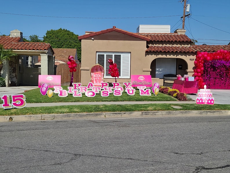May include: Pink and gold Happy Birthday Blossom yard signs with a pink and gold 15 sign, pink and gold balloons, and a pink and gold cake. The signs are on a green lawn in front of a house.