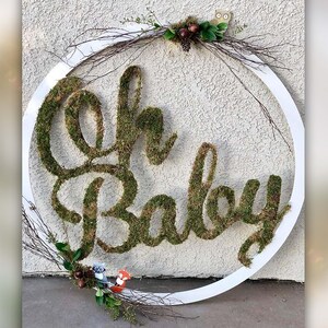 May include: A white circular wreath with the words "Oh Baby" spelled out in moss. The wreath is decorated with small woodland creatures and greenery.
