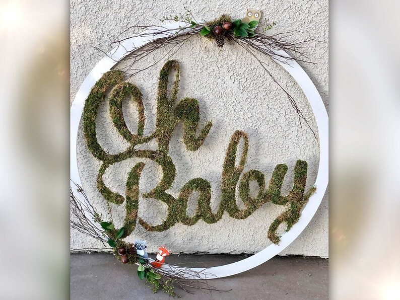 May include: A white circular wreath with the words "Oh Baby" spelled out in moss. The wreath is decorated with small woodland creatures and greenery.