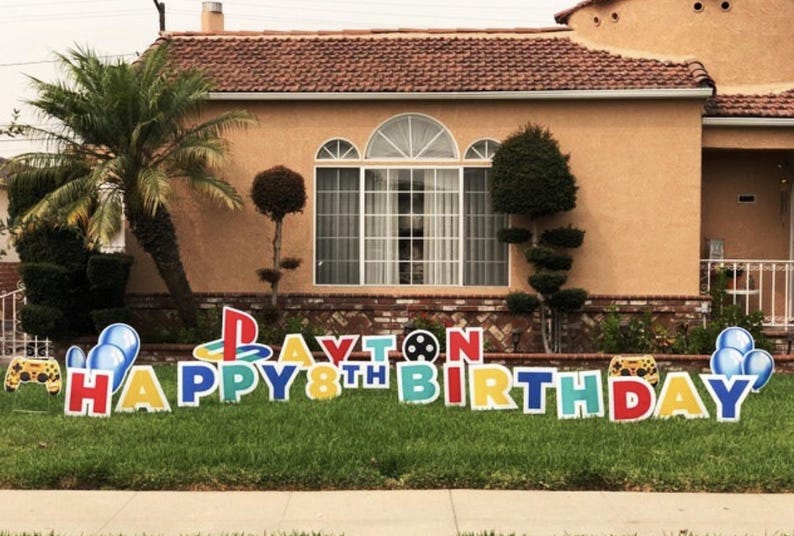 May include: A colorful yard sign with the words "Happy 8th Birthday" and the name "Payton" in large letters. The sign also features a video game controller, a PlayStation logo, and balloons.