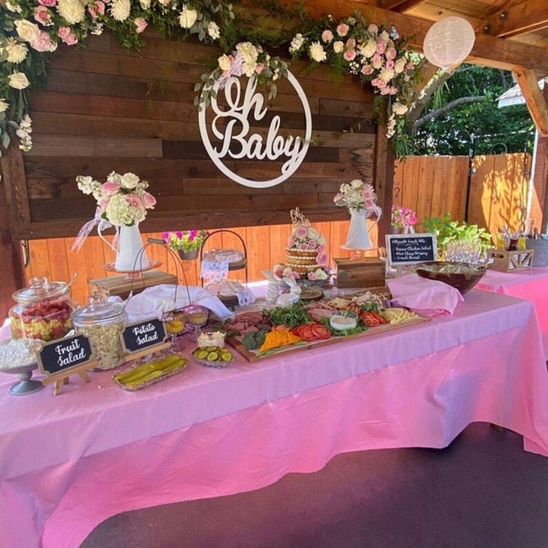 May include: A pink table with a variety of food on it, including a fruit salad, potato salad, and a charcuterie board. There is a sign that says "Oh Baby" in the background.