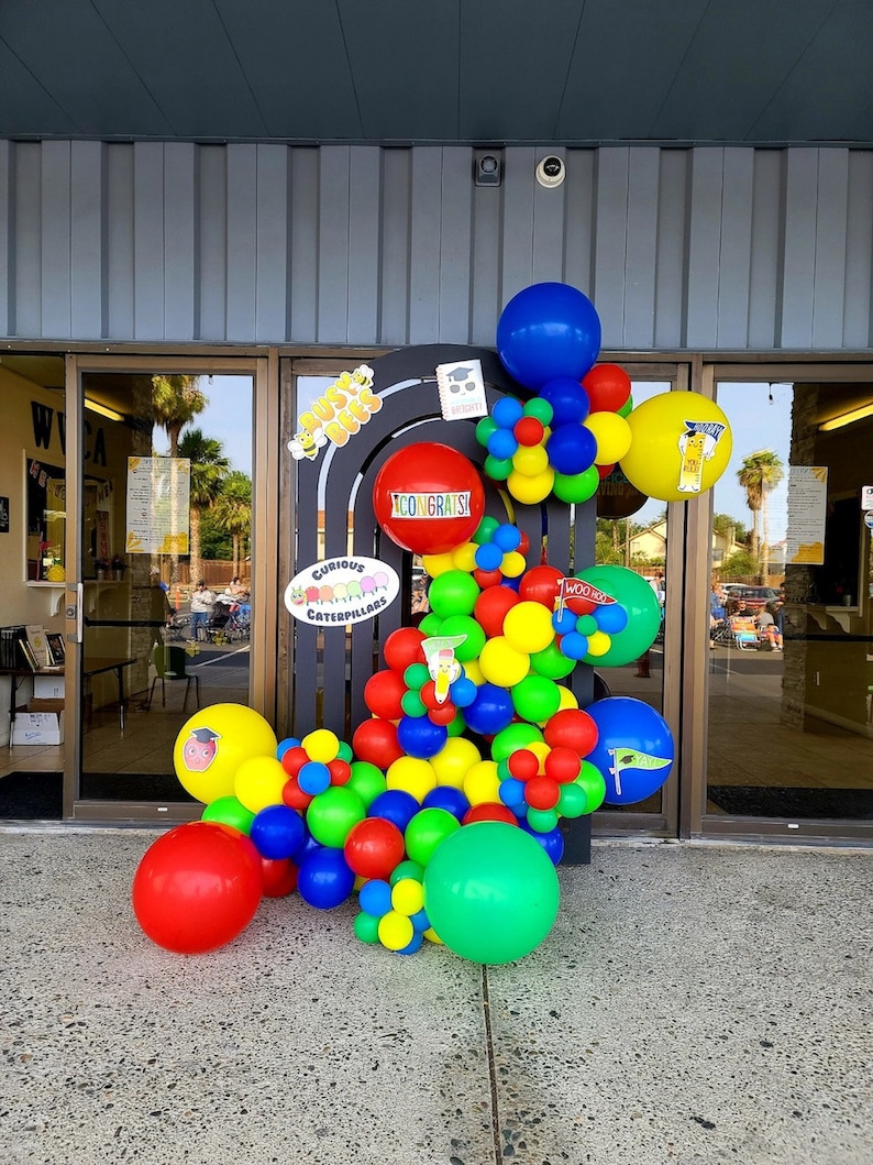 Pu&ograve; includere: Un arco di palloncini colorati decorato con le parole "Curious Caterpillars" e "Woo Hoo!" L'arco &egrave; fatto di palloncini rossi, gialli, blu e verdi. Ci sono anche alcuni piccoli palloncini bianchi con punti neri.