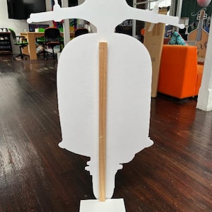 May include: A white scooter-shaped cutout with handlebars, a seat, and a base. A wooden support beam runs vertically down the centre, connecting the scooter to a rectangular base. The scooter is positioned in an office setting with wooden flooring.