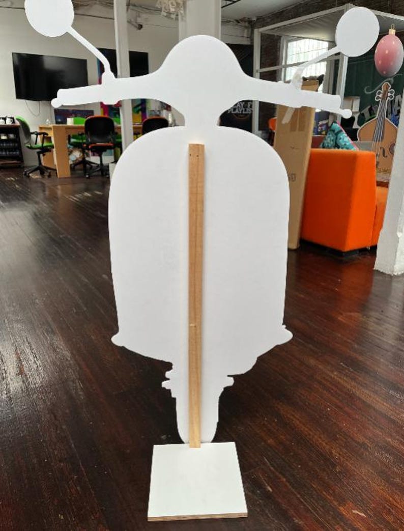 May include: A white scooter-shaped cutout with handlebars, a seat, and a base. A wooden support beam runs vertically down the centre, connecting the scooter to a rectangular base. The scooter is positioned in an office setting with wooden flooring.