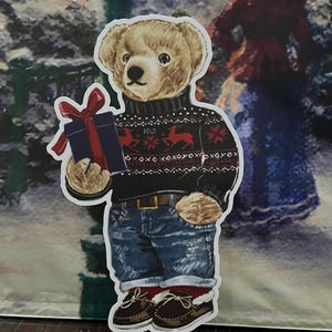 May include: A cutout of a teddy bear wearing a black sweater with a red and white pattern, blue jeans, and brown shoes. The bear holds a blue gift box with a red bow. The sweater has the word "POLO" on it.