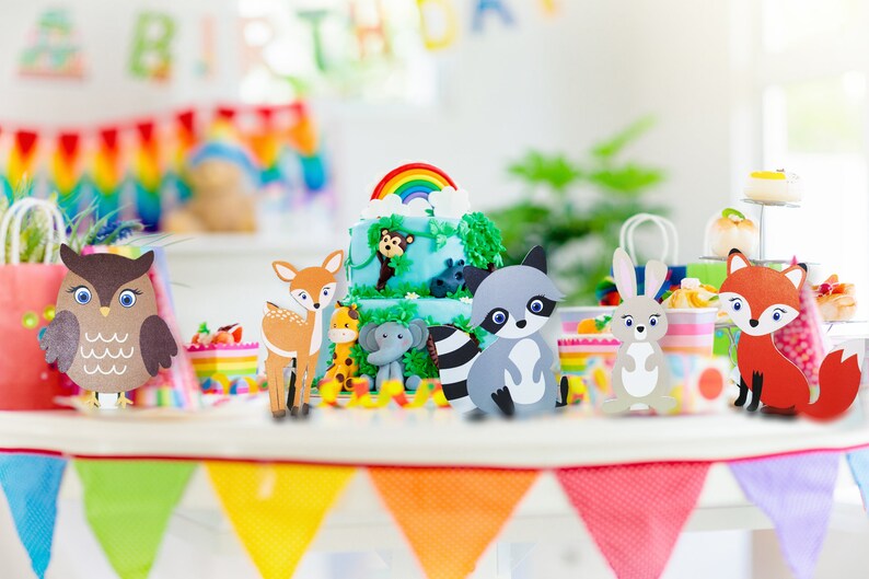 May include: A set of woodland animal cake toppers for a birthday party. The toppers are made of cardboard and feature a raccoon, owl, deer, fox, rabbit, elephant, giraffe, and monkey. The toppers are arranged on a table with a cake in the background.