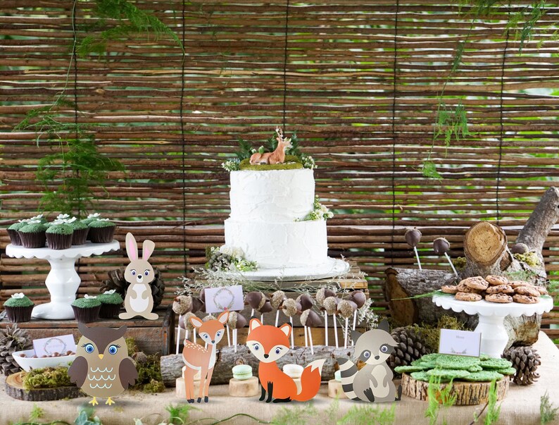May include: A woodland-themed party table setting with a white two-tier cake decorated with a deer figurine. The table is decorated with cupcakes, cookies, and various animal figurines, including a fox, owl, deer, rabbit, and raccoon.