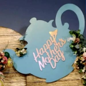 May include: A teal blue teapot shaped sign with the words "Happy Mother's Day" in pink lettering. The sign is decorated with flowers and greenery.