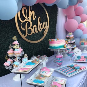 May include: A gender reveal party dessert table with a blue and pink balloon arch, a cake with blue and pink frosting, cupcakes, cookies, and other treats. The table is decorated with a gold "Oh Baby" sign.