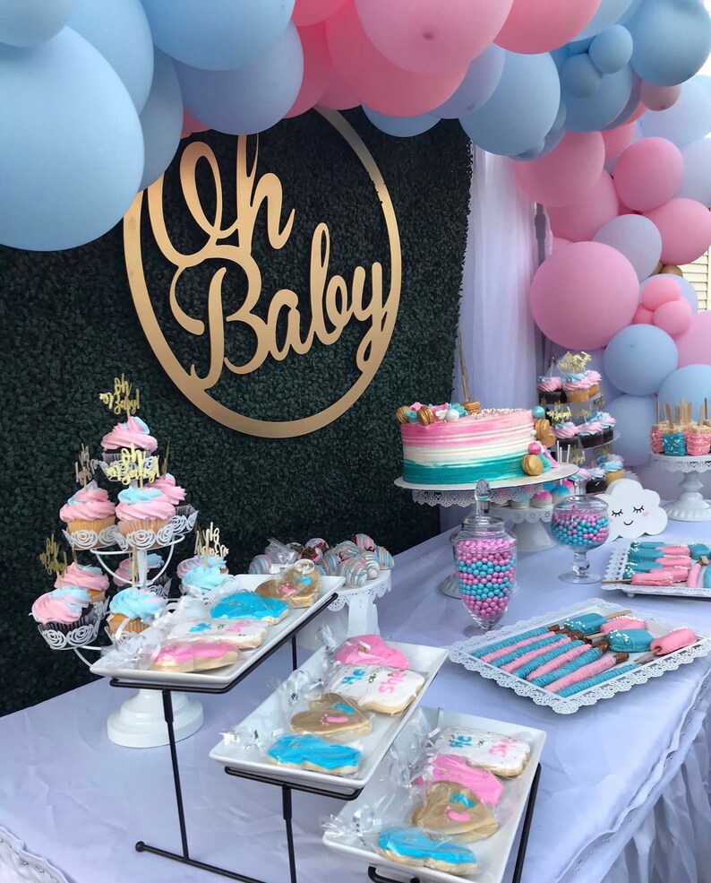 May include: A gender reveal party dessert table with a blue and pink balloon arch, a cake with blue and pink frosting, cupcakes, cookies, and other treats. The table is decorated with a gold "Oh Baby" sign.