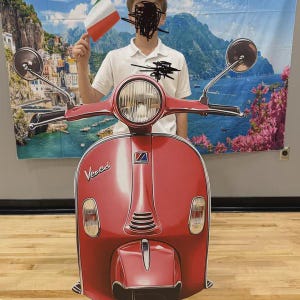 May include: A red Vespa scooter cutout with a large headlight, side mirrors, and the word "Vespa" on the front. The scooter is in front of a person holding an Italian flag. The background features a scenic mural.