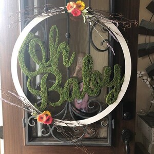 May include: A white wreath with a green moss sign that says "Oh Baby" and a floral arrangement with pink and yellow flowers.