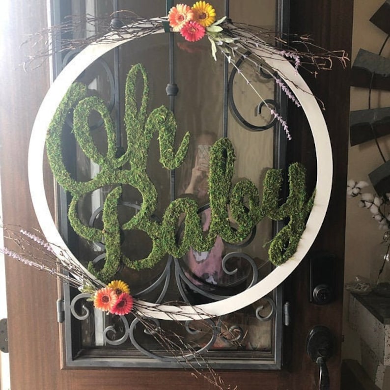 May include: A white wreath with a green moss sign that says "Oh Baby" and a floral arrangement with pink and yellow flowers.