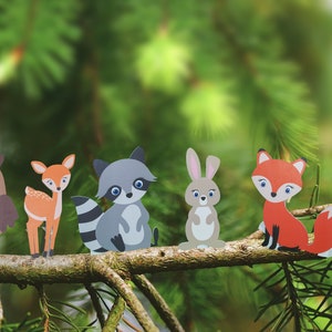 May include: Five paper cutouts of woodland animals on a pine branch. The animals are an owl, a deer, a raccoon, a rabbit, and a fox. The animals are all facing forward and are arranged in a line.