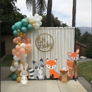 May include: A white backdrop with a gold circle sign that says "Oh Baby" and a balloon arch with peach, white, and teal balloons. There are five woodland animal cutouts: a deer, a fox, a raccoon, an owl, and a rabbit.