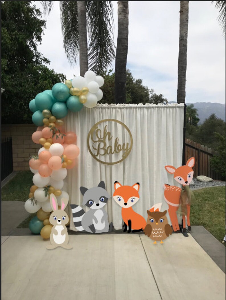May include: A white backdrop with a gold circle sign that says "Oh Baby" and a balloon arch with peach, white, and teal balloons. There are five woodland animal cutouts: a deer, a fox, a raccoon, an owl, and a rabbit.