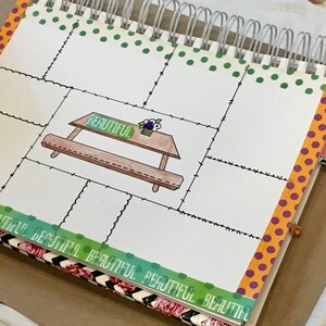 Puede incluir: Un diario blanco con un borde de cinta de lunares verdes, naranja y morado. El diario tiene una encuadernación en espiral y una página con una ilustración dibujada a mano de una mesa de picnic con la palabra "Beautiful" escrita en ella. La página está dividida en secciones con un borde de línea negra ondulada. Una pestaña rosa con el número "31/01" está unida al diario.
