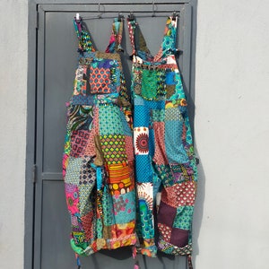 May include: Two pairs of colorful patchwork overalls hanging on a hanger against a gray wall. The overalls are made of different fabrics with various patterns and colors, including blue, green, red, yellow, and orange.