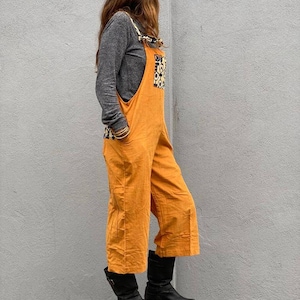 May include: A woman wearing a mustard yellow overall with a black and white patterned patch on the front. She is wearing black boots and standing on a rock.