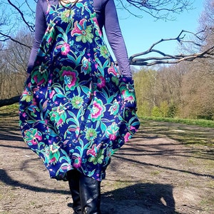 May include: A woman wearing a blue and green floral print dress with a long-sleeved purple shirt underneath. She is wearing black boots and standing in a grassy area.