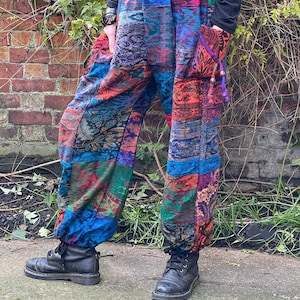 May include: Multi-coloured patchwork trousers with an elasticated waist and pockets. The trousers feature a variety of patterns and are gathered at the ankles. The person is wearing black boots. The colours include blue, red, and green.
