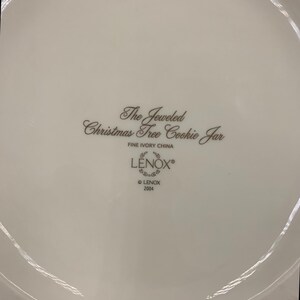 May include: The bottom of a white ceramic cookie jar with the words "The Jeweled Christmas Tree Cookie Jar" and "LENOX" in brown lettering. The jar is made of fine ivory china and was made in 2004.