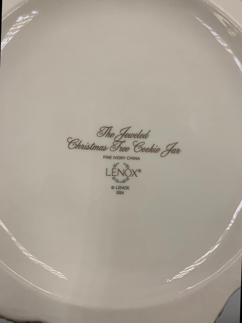 May include: The bottom of a white ceramic cookie jar with the words "The Jeweled Christmas Tree Cookie Jar" and "LENOX" in brown lettering. The jar is made of fine ivory china and was made in 2004.