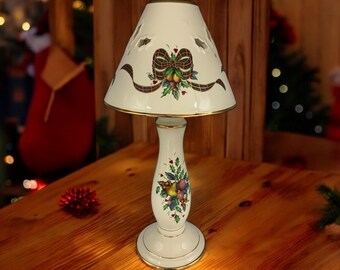 May include: A white ceramic table lamp with a gold trim and a festive design. The lampshade features a red bow, green holly leaves, and red berries. The base of the lamp has a similar design with a gold trim.