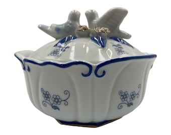 May include: A white porcelain trinket dish with a blue and white floral design. The lid features two white porcelain birds with gold accents.