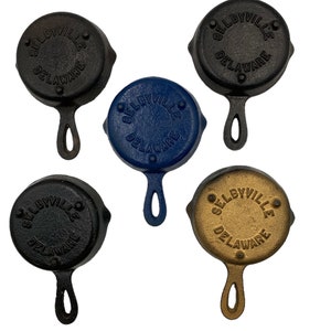 Puede incluir: Cuatro peque&ntilde;as sartenes de hierro fundido con asas. Cada sart&eacute;n es de un color diferente: negro, azul, dorado y negro. Las sartenes est&aacute;n todas estampadas con las palabras "Selbyville, Delaware".