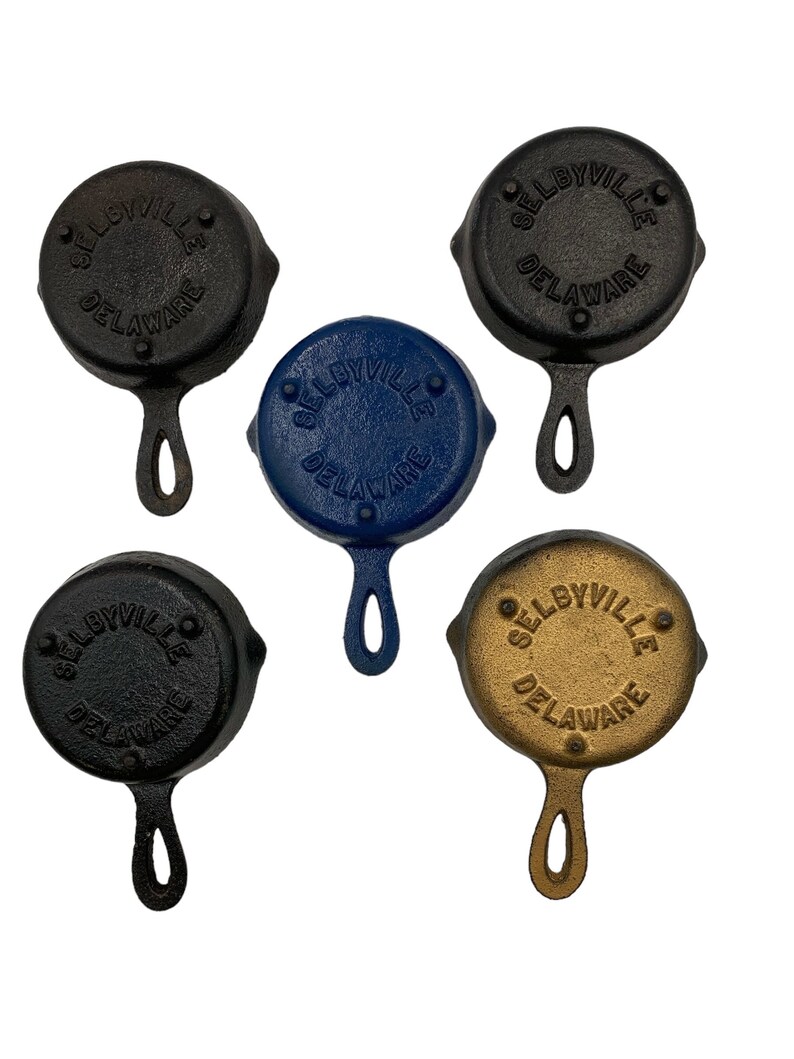 Puede incluir: Cuatro peque&ntilde;as sartenes de hierro fundido con asas. Cada sart&eacute;n es de un color diferente: negro, azul, dorado y negro. Las sartenes est&aacute;n todas estampadas con las palabras "Selbyville, Delaware".