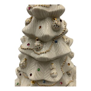 May include: A white ceramic Christmas tree with gold trim and colorful ornaments. The tree is decorated with a gold chain and small, colorful gems.