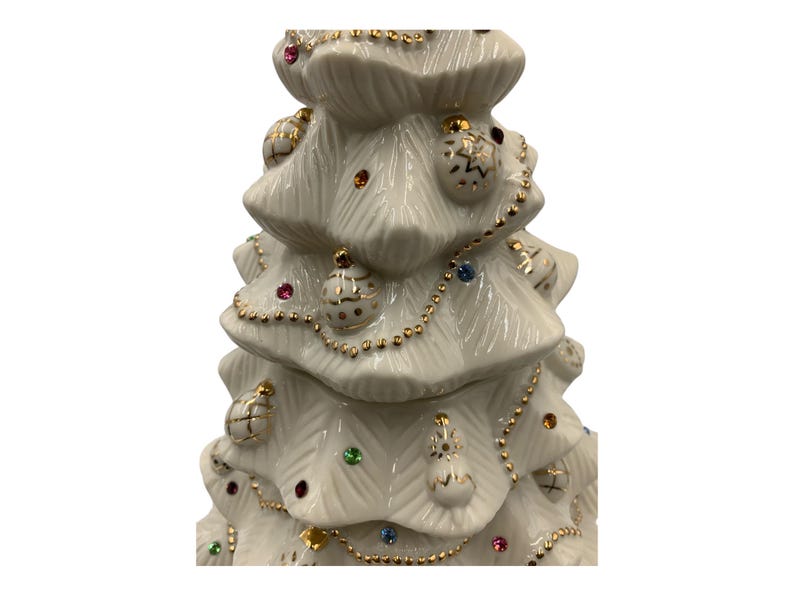 May include: A white ceramic Christmas tree with gold trim and colorful ornaments. The tree is decorated with a gold chain and small, colorful gems.