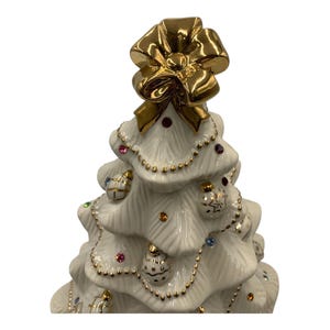 May include: A white porcelain Christmas tree figurine with a gold bow on top and gold trim. The tree is decorated with colorful jewels and gold ornaments.