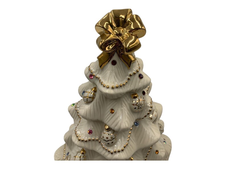 May include: A white porcelain Christmas tree figurine with a gold bow on top and gold trim. The tree is decorated with colorful jewels and gold ornaments.