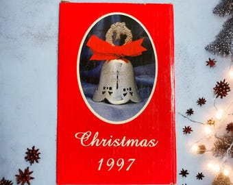 May include: A red box with a silver bell ornament inside. The bell has a red ribbon and a wreath on top. The box says "Christmas 1997" on the front.