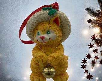 May include: A yellow cat wearing a white and red Santa hat with a green holly sprig ornament. The cat is holding a gold bell in its paws.