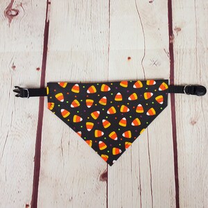 K&ouml;nnte beinhalten: Schwarzes Hundehalstuch mit einem Candy-Corn-Muster. Das dreieckige Halstuch hat einen schwarzen Hintergrund mit orangefarbenen, gelben und wei&szlig;en Candy-Corn-Formen und bunten Punkten. Es hat ein verstellbares schwarzes Halsband mit einer Kunststoffschnalle.