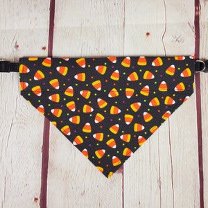 K&ouml;nnte beinhalten: Ein schwarzes Hunde-Bandana mit einem Candy-Corn-Muster. Das Bandana ist dreieckig und hat ein schwarzes, verstellbares Halsband. Das Candy-Corn-Design zeigt orange, gelbe und wei&szlig;e Candy-Corn-Formen mit kleinen bunten Punkten.