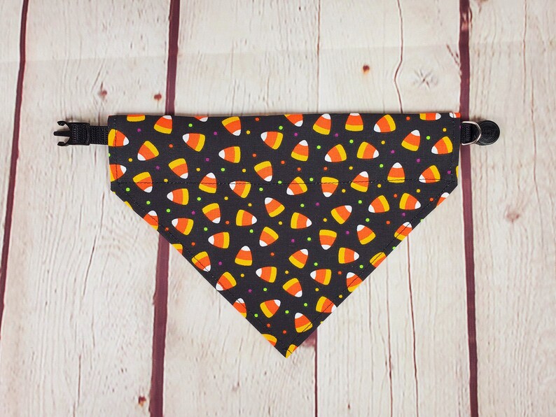 K&ouml;nnte beinhalten: Ein schwarzes Hunde-Bandana mit einem Candy-Corn-Muster. Das Bandana ist dreieckig und hat ein schwarzes, verstellbares Halsband. Das Candy-Corn-Design zeigt orange, gelbe und wei&szlig;e Candy-Corn-Formen mit kleinen bunten Punkten.