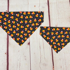 K&ouml;nnte beinhalten: Zwei schwarze, dreieckige Hunde-Bandanas mit einem sich wiederholenden Muster aus Candy Corn und bunten Punkten. Die Bandanas sind aus Stoff und f&uuml;r das Tragen um den Hals eines Hundes konzipiert. Halloween-Thema.