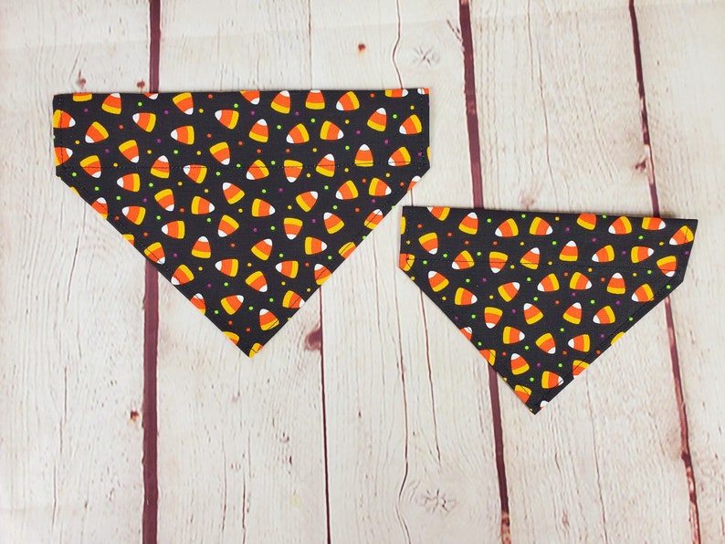 K&ouml;nnte beinhalten: Zwei schwarze, dreieckige Hunde-Bandanas mit einem sich wiederholenden Muster aus Candy Corn und bunten Punkten. Die Bandanas sind aus Stoff und f&uuml;r das Tragen um den Hals eines Hundes konzipiert. Halloween-Thema.