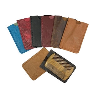 Könnte beinhalten: Eine Sammlung von Leder-Kartenhaltern in verschiedenen Farben, darunter Blau, Rot, Schwarz und Braun. Ein Holzkamm ist sichtbar, teilweise in einem schwarzen Lederetui und einem hellbraunen Lederetui eingesetzt. Die Kartenhalter sind fächerförmig angeordnet.