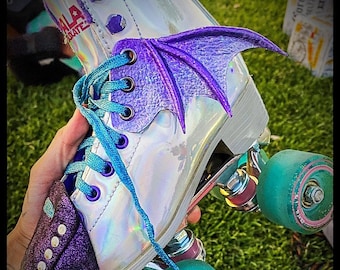 May include: A pair of iridescent white roller skates with purple and blue accents. The skates have a purple and blue wing design on the front and a purple and blue toe stop. The wheels are green with pink accents.